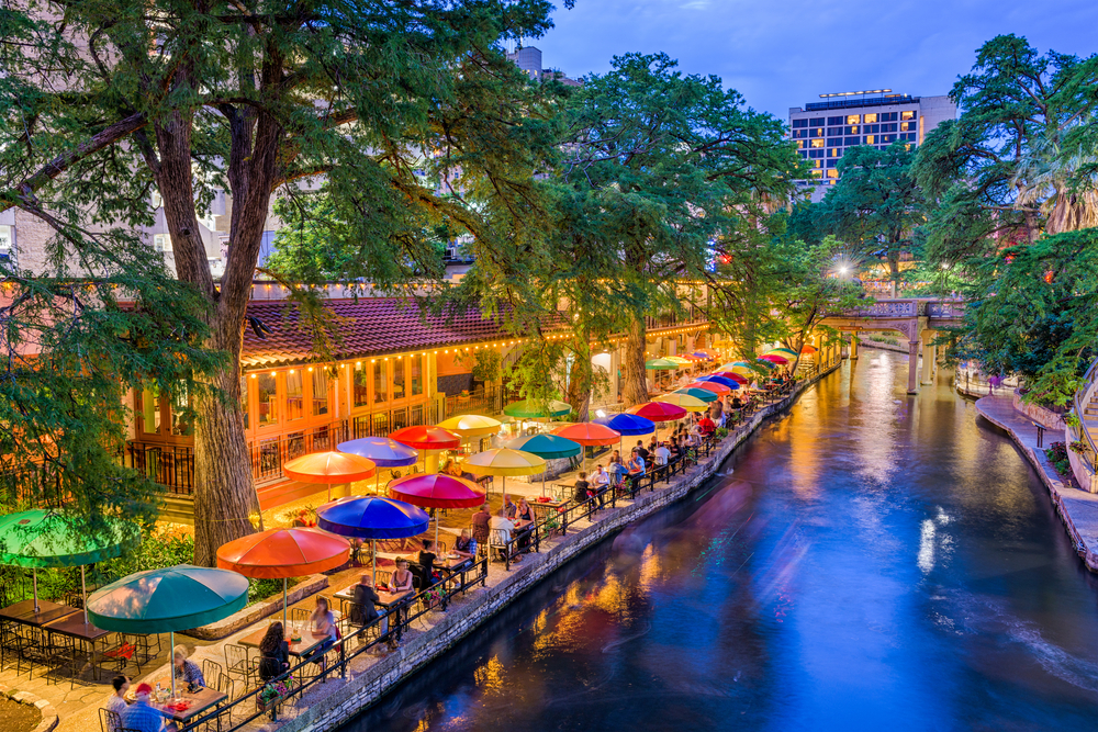 people sitting on tables under colorful umbrellas beside a river san antonio riverwalk restaurants