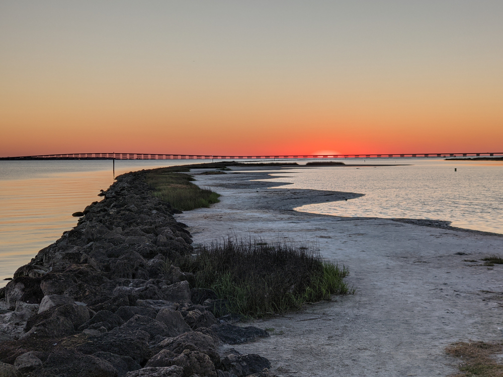 rocks and sandy path surrounded by water with a pier and orange sky in the background