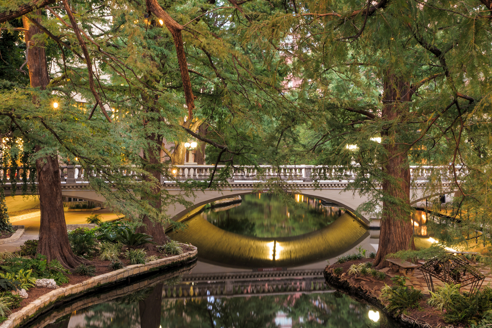 silver colored bridge surrounded by trees and passing over a river san antonio riverwalk