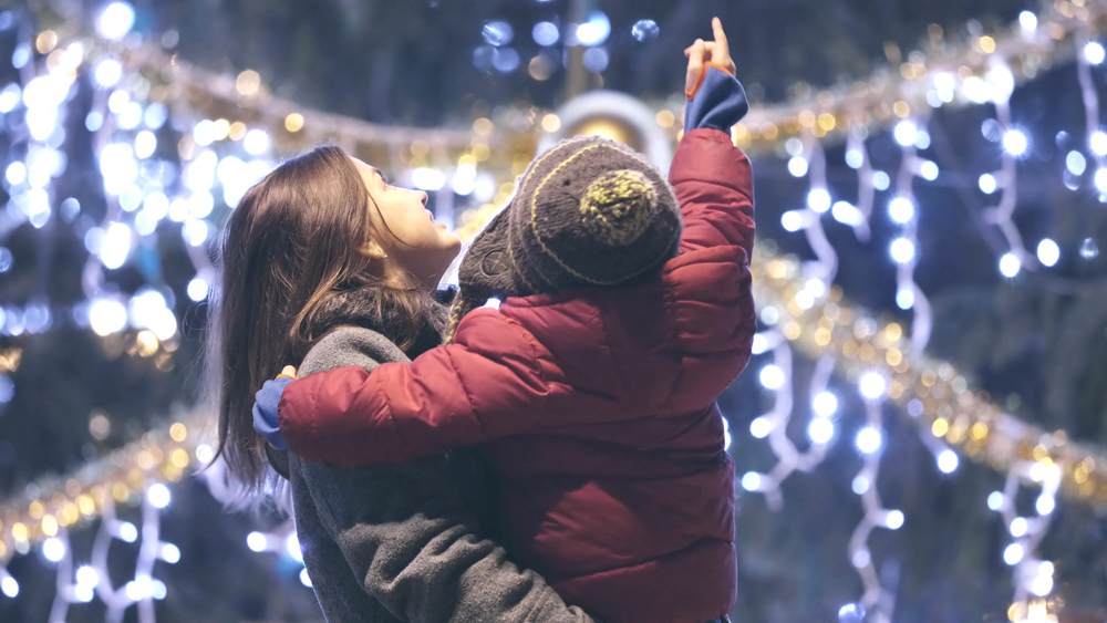 A photo of a mother holding her child as they point up at Christmas lights glowing in the background.