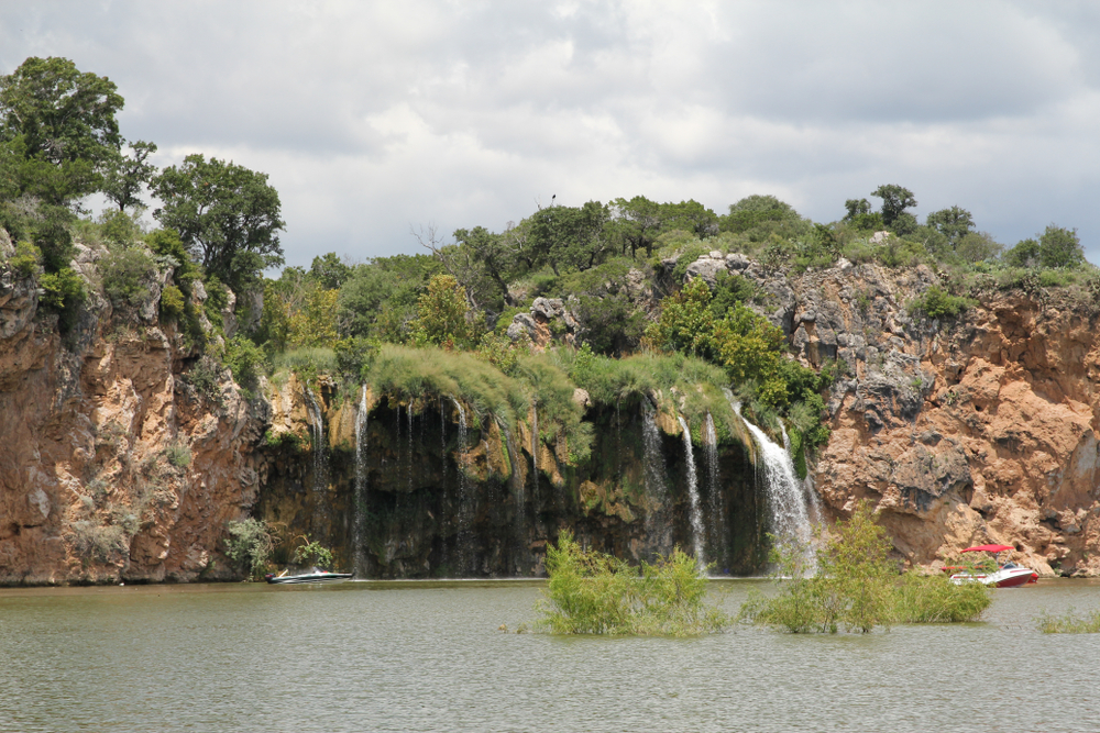 two boats appear small in the water up against a waterfall with reddish rocky walls, and green vegetation in the water.