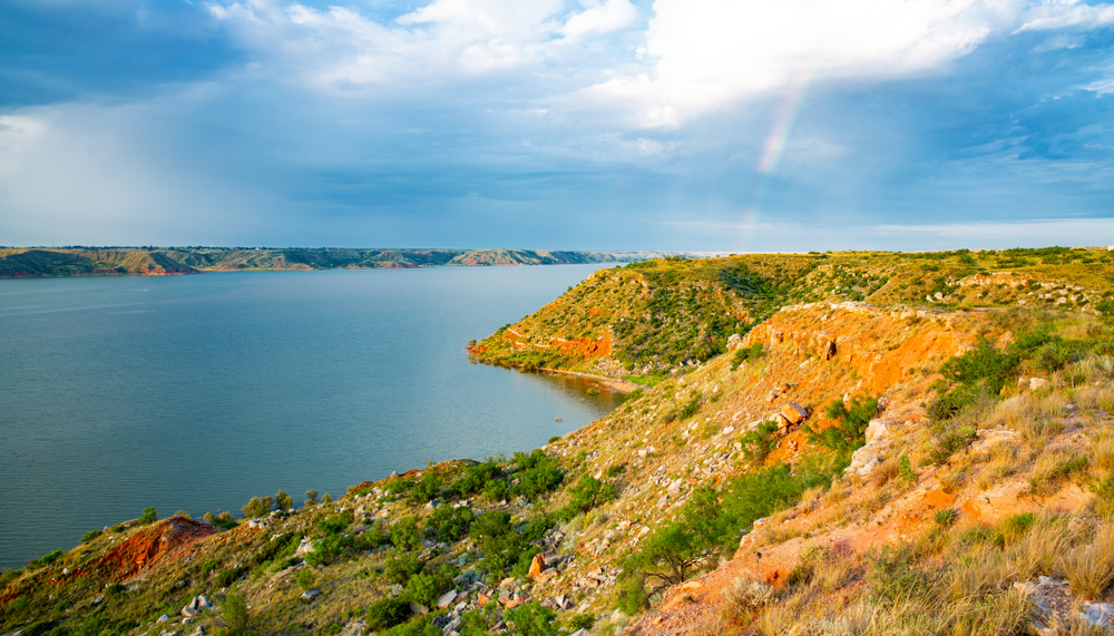 Orangish rocky coastline in the foreground blue lake waters in the background, far away coastline