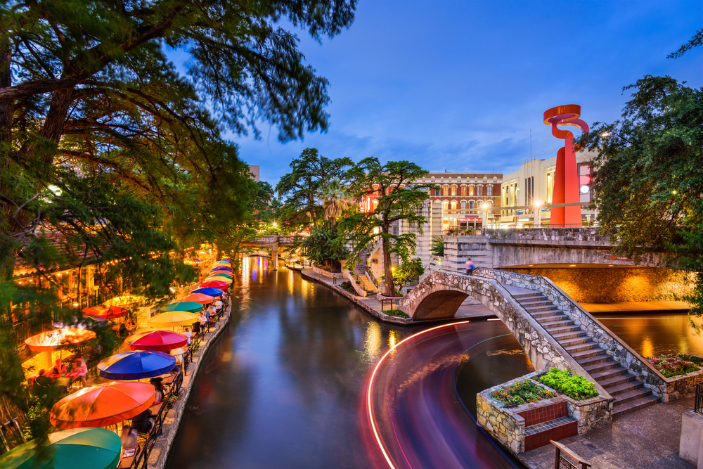 An evening photo of the Riverwalk in San Antonio. Tables with brightly colored umbrellas line the edge of the river.