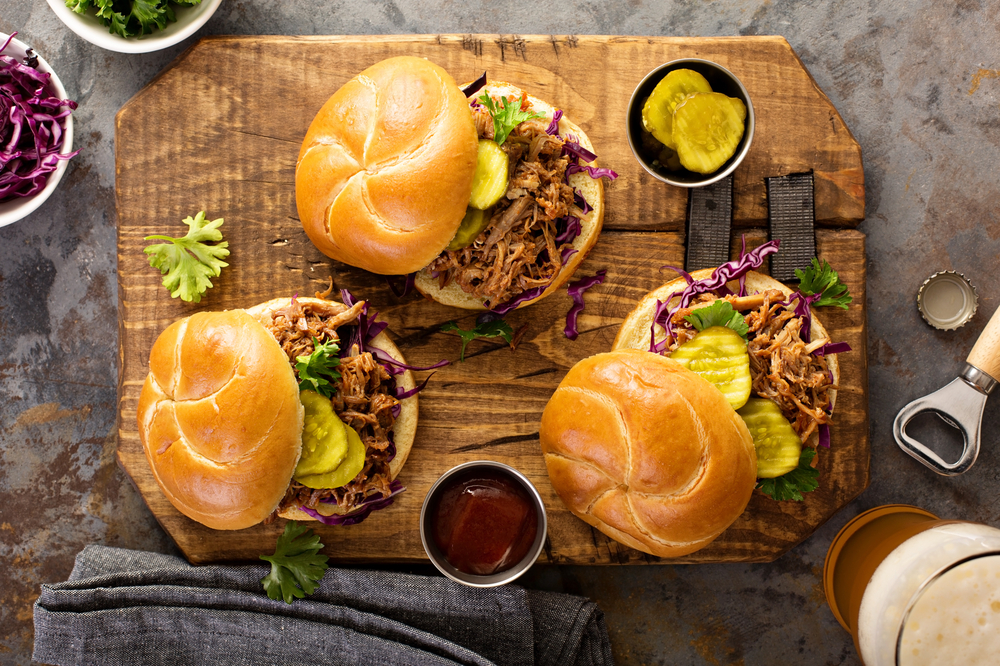 A cutting board holding three pulled pork sandwiches with pickles and bbq sauce. Get one just like it at the Big Bib BBQ, a black-owned restaurant in San Antonio
