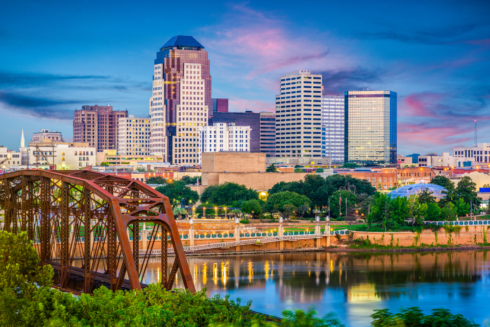 A rusty bridge sits in front of the lights of the Shreveport city skyline as they reflect off a river at twilight. 