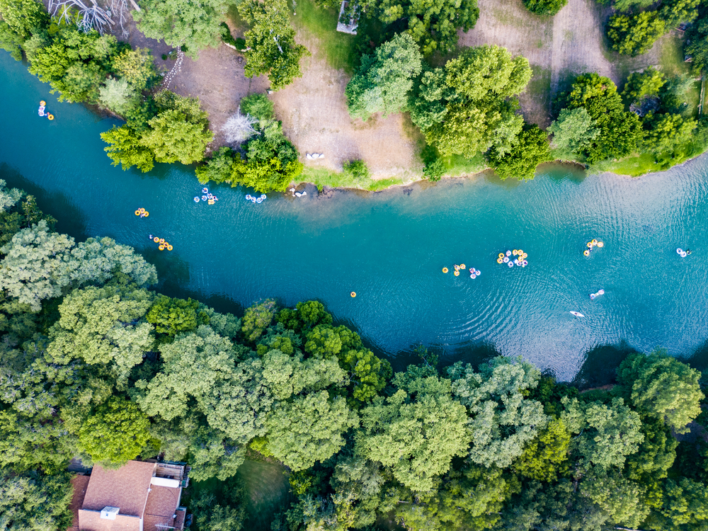 An aerial shot of the people tubing the crisp blue Guadalupe River that cuts through the center of a forest. 