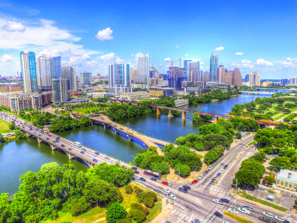 An aerial view of the Austin skyline. There are bridges going over a large river and you can see cars on them. There are lots of tall buildings, green space and trees near the river, and the sky is very blue. 