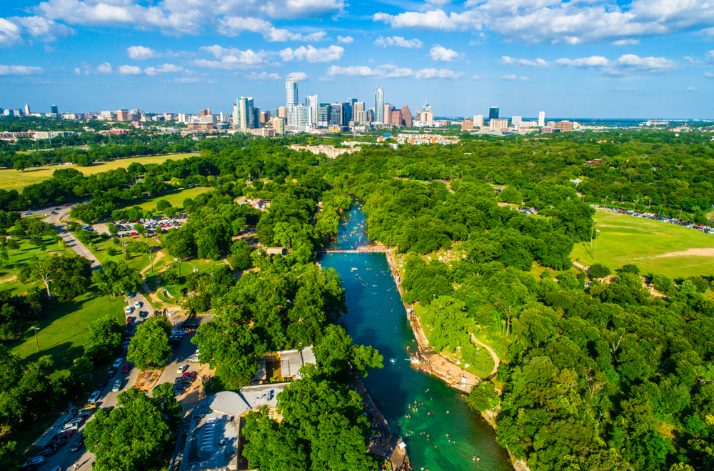 An aerial view of a large long pool that is fed by a natural spring. The pool is a long rectangle that is in a grassy park. There are trees surrounding the pool. The water is a beautiful blue and you can see people swimming in it. In the distance you can see the Austin skyline. One of the best Texas day trips.