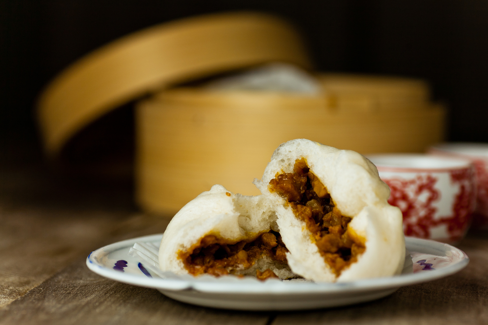 A small plate with two Asian steamed bbq pork rolls. you can see the inside of the rolls where there is meaty filling. In the background you can see an Asian steaming basket and two red and white ceramic cups. 