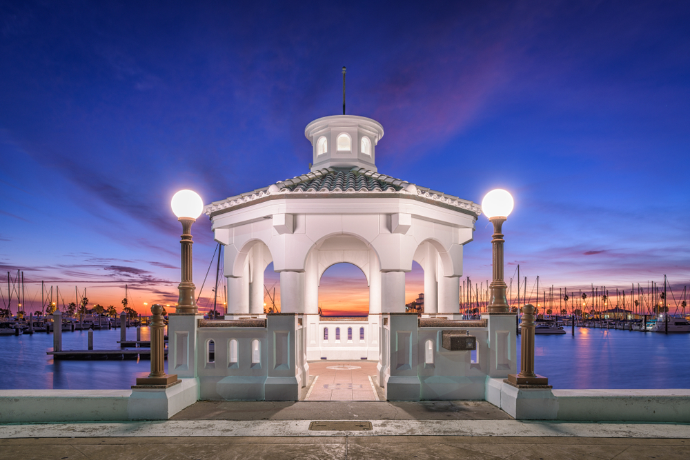A beautiful white gazebo lit up at night with a sunset sky and docked boats in the background 