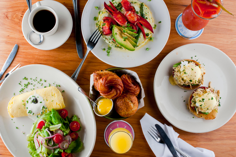 A spread of classic brunch foods. There is eggs benedict, a omelette, pastries, grits with crab claws and sliced avocado, and glasses with juice and coffee. 