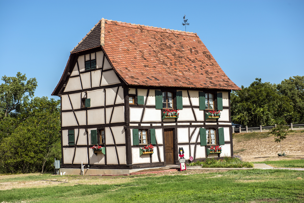 An antique home that looks like a classic Swiss Alps style home. It is white stucco with dark wood support beams on it. The windows have dark green shutters with plant boxes with red flowers under them. Around the house is a grassy yard and some trees. 