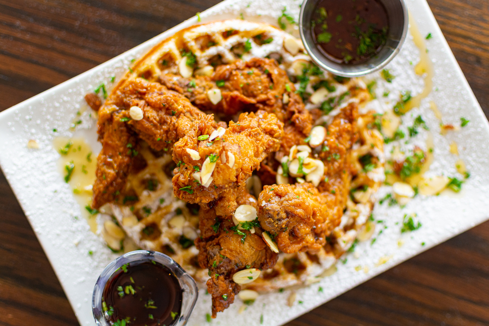 A white plate from a restaurant in Amarillo with a large waffle topped with fried chicken. There are slivers of nuts, some type of green herb, and powdered sugar. There are two bowls of syrup on the plate. 