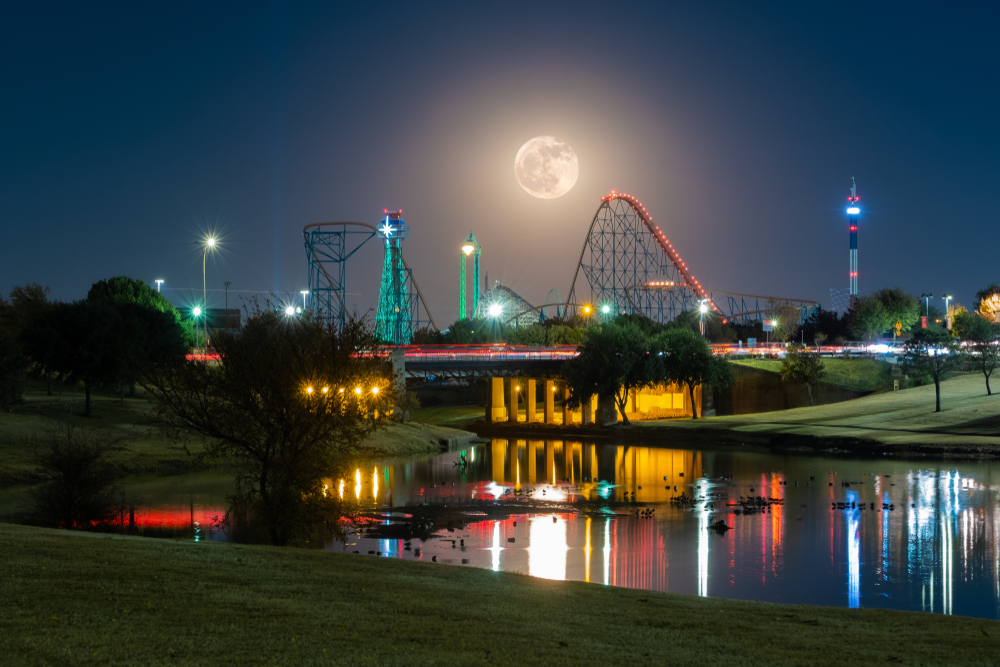 Moonlit amusement park with  skyline of roller-coasters with road and pond in front