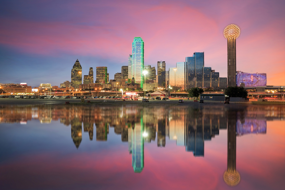 The Dallas skyline at twilight from the view of the river. The skyline is all lit up and the sky is mostly pink and purple with some blue. The sky and the skyline is reflected in the river. 