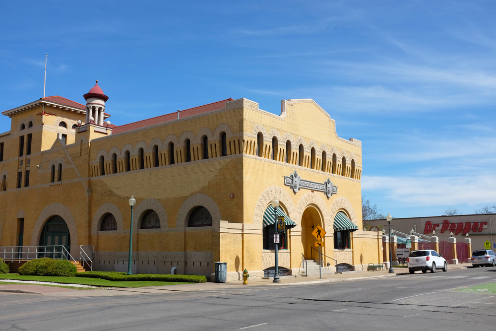 The exterior of a 20th century building. It is a creamy color and has arched windows all along the top. There is an arched doorway and behind it you can see a building that says 'Dr. Pepper'. It is one of the best day trips from Dallas. 