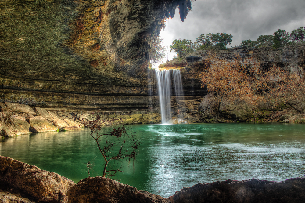 A dripping waterfall over a rocky cliff. It is falling into a greenish pool of water and there is a large rock overhang around the pool. There are some dead trees near the pool. 