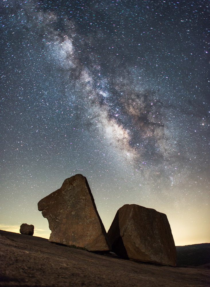 An image of two large uniquely shaped rocks with the Milky Way in the sky behind them. The rocks are somewhat in shadow. 