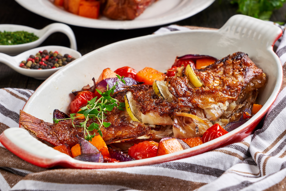 A fish in a deep white and red dish. It is cooked and crispy on the outside. You can see lemon slices cut and placed into the side of the fish. In the dish there is also cooked vegetables like potato, onion, squash, and tomato. There is fresh oregano on it and the dish is wrapped in a stripped kitchen towel. 