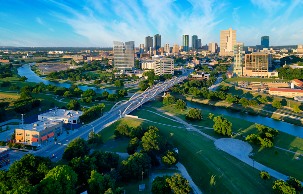 An aerial view of Forth Worth, a city just outside of Dallas. There is a river running through the city, a bridge with arches on it, and a typical city skyline. Near the river there are a lot of grassy areas and trees. It is a sunny day. 