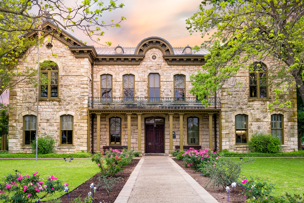 An old stone building that is architecturally influenced by antique German building. In front of it is a walkway that has flower beds with pink flowers on either side and then a grassy lawn. You can also see a few trees. 