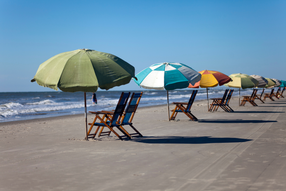 A group of beach chairs and different colored umbrellas all lined up. They are sitting on the sandy shore of Galveston Beach, one of the best Texas day trips. The chairs are facing the ocean and you can see waves crashing and someone standing in the water. 