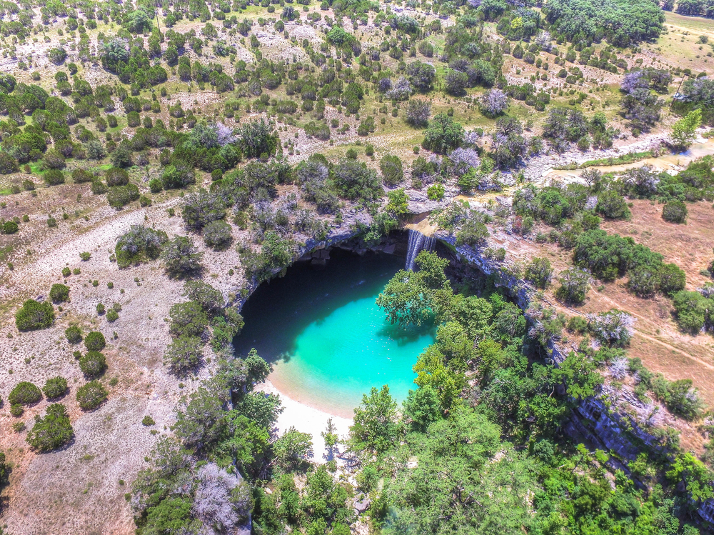 An aerial view of the Hamilton Pool, one of the best Texas day trips. You can see a pool of crystal blue water inside what looks like a sunken cave or crater. Around it is a desert landscape with shrubs and some grass. 