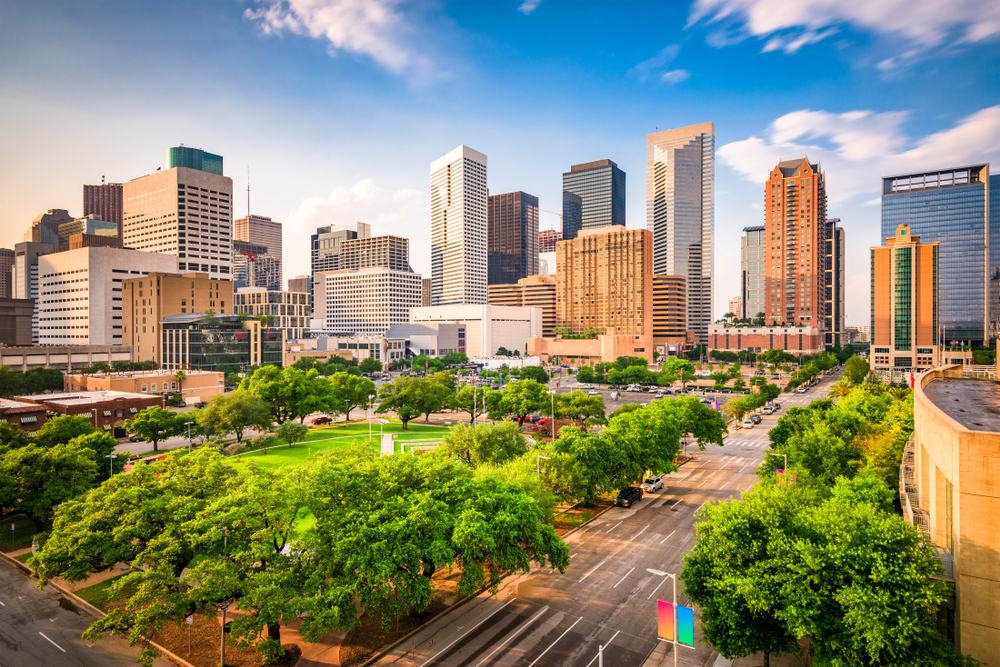 The skyline of Houston Texas. There are lots of skyscrapers. You can also see a large park with trees all around it and a large green space in the middle. It is a sunny day with a few clouds in the sky. One of the best day trips from Dallas. 