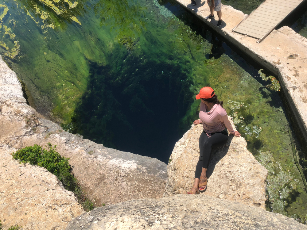 A woman wearing black leggings, a blush colored shirt, and a red hat sitting on a rock. The rock is hanging over one of the underwater caves in Jacobs Well. The cave is a large hole that descends into total darkness. The water is crystal clear and the rocks have green moss growing on them. 