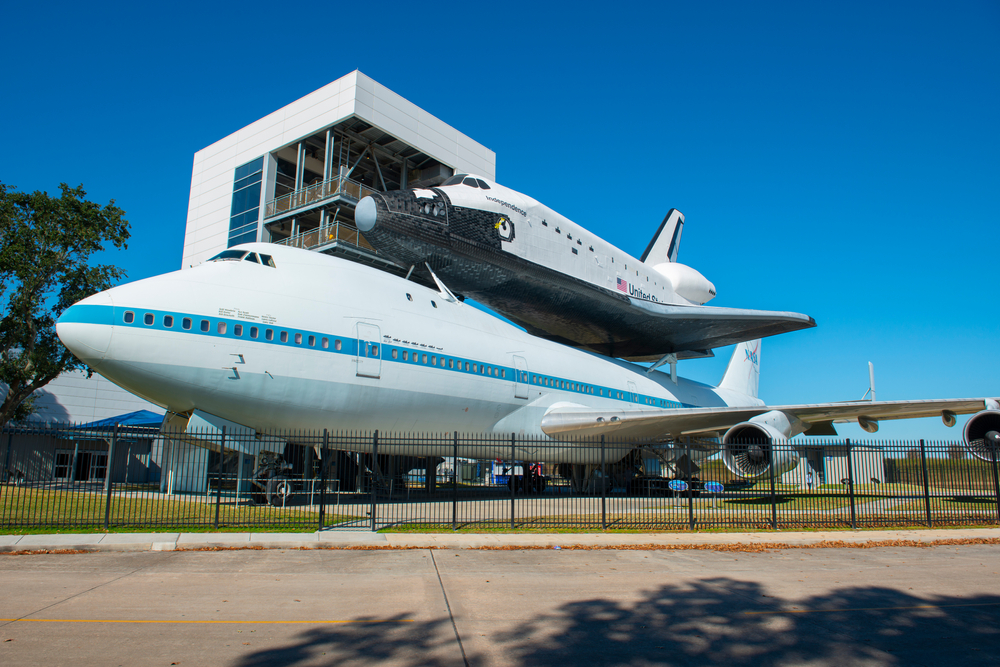 A NASA space shuttle perched on a large airplane. There is a black fence surrounding it and behind it you can see a square building with decks on it. It is one of the best Texas day trips. 