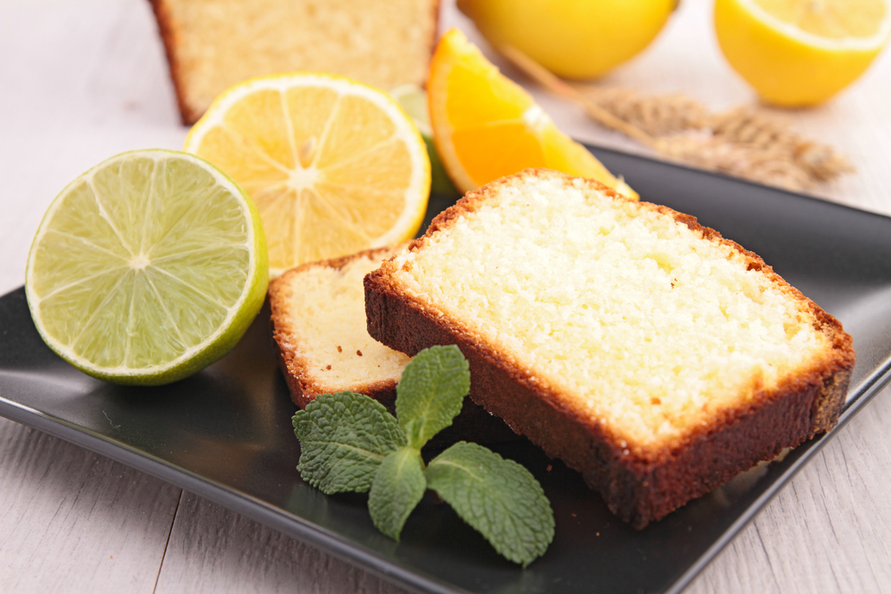 Two slices of lemon pound cake on a black ceramic plate. On the plate there are also slices of lemon, lime, and orange. There is also a sprig of fresh mint. 