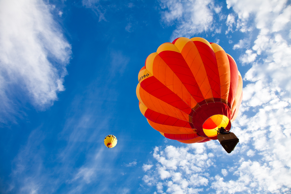 Looking up at two hot air balloons in the sky. The sky is very blue with some scattered clouds. The balloons are chevron patterned in yellow, orange, and red. 