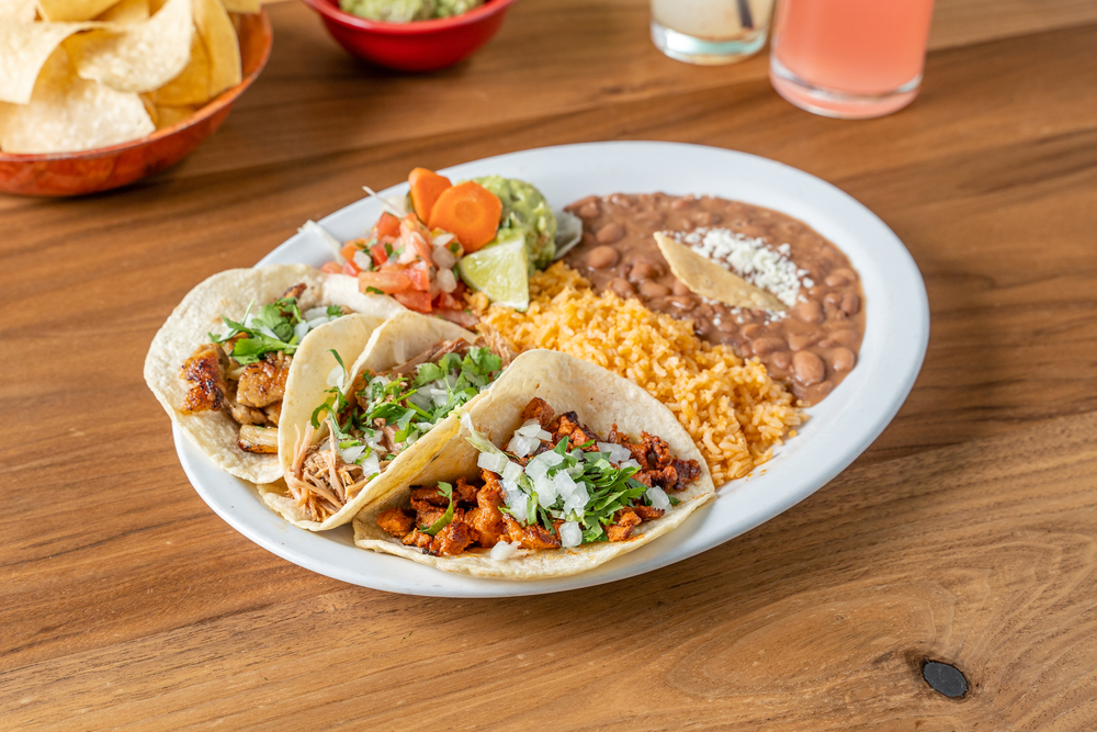 A white plate on a wooden table with a traditional authentic Mexican meal on it. There are refried beans, saffron rice, and three tacos with different meats in each of them. 