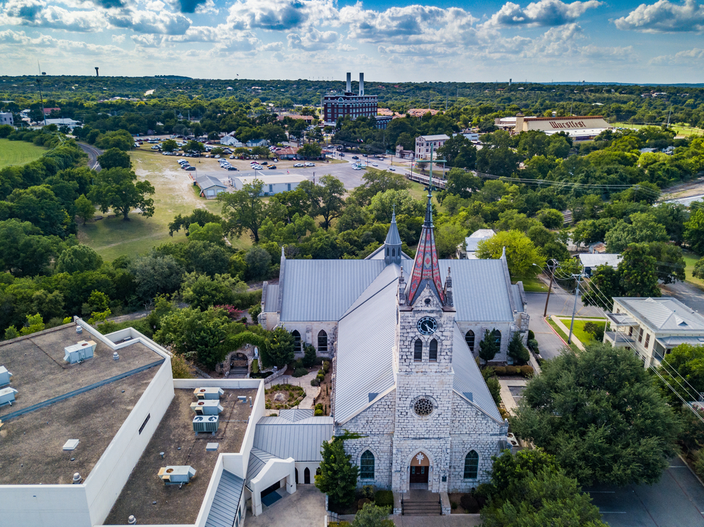 An aerial view of an old town in Texas. You can see a church steeple with white, black, and white patterned shingles and other antique buildings. The town has a lot of trees with green leaves. Just a little in the distance there appears to be an old factory building with two smoke stacks. 