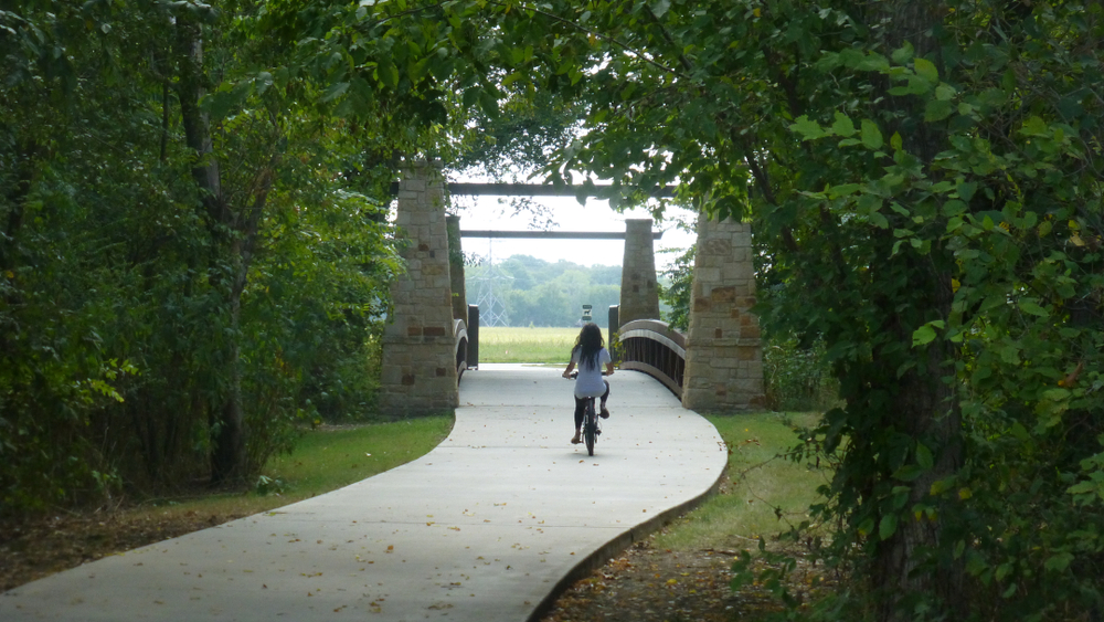 A woman with long hair riding a bike on a pathed trail. The trail is surrounded by green trees and grass. In the distance there is a bridge and you can see a field with trees in it. It is one of the best day trips from Dallas. 