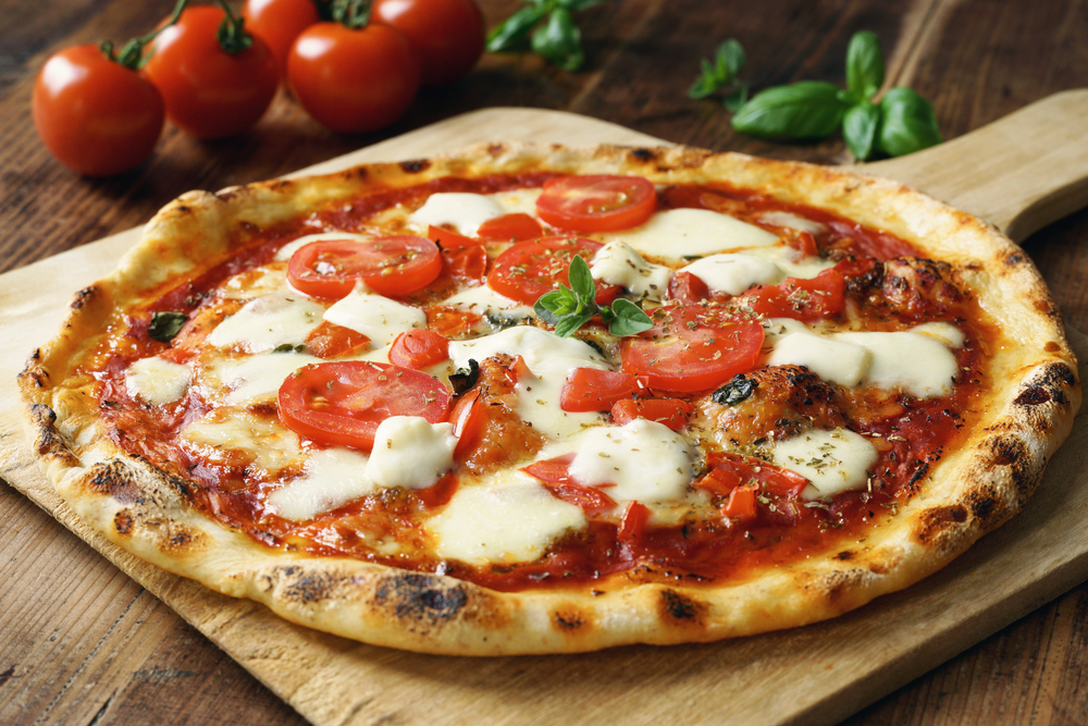 A large Margherita pizza on a wooden paddle. There is also fresh tomato and basil on the cooked pizza. It is on a wooden table and in the background you can see some tomatoes and fresh basil. 