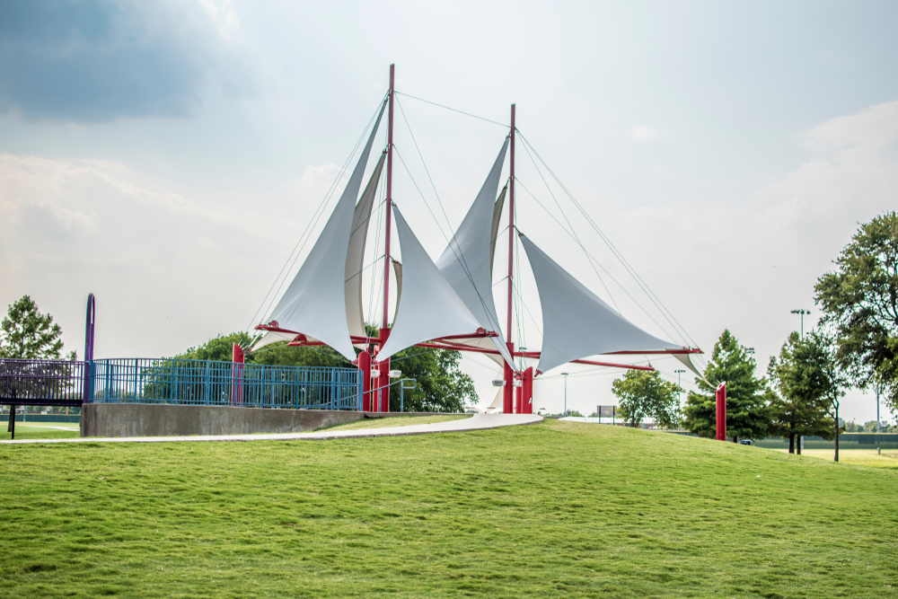 A sailboat sculpture that is just the sails. The sails are white and there are red metal bars connecting the sails. The sculpture is on a grassy lawn and has a sidewalk leading up to it. There some trees nearby.