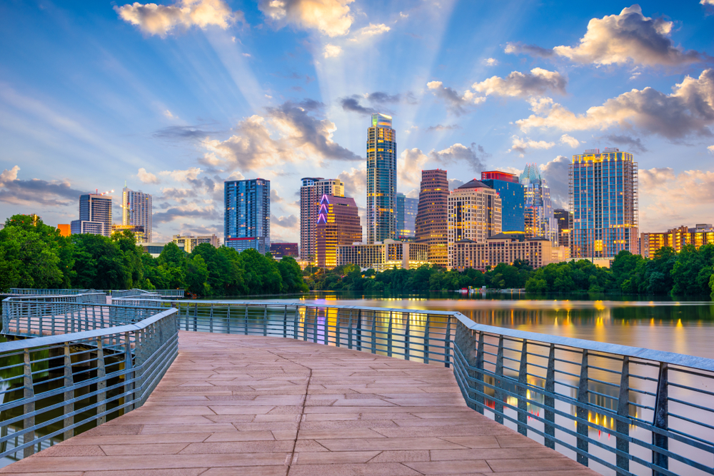 Looking at the Austin city skyline from a paved walkway that crosses over the river. It is twilight so the sun is starting to set and the buildings are all lit up. You can see lights reflecting in the river. 