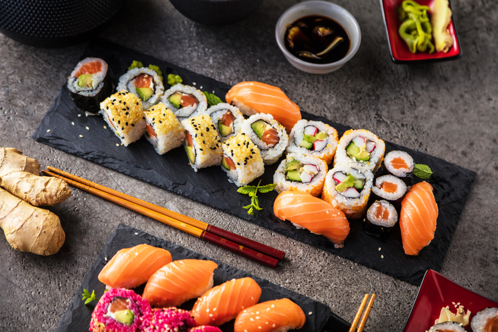 Two black slate trays with various sushi rolls on them. Some of them are covered in rice, have sesame seeds on them, or are covered in nori. There is also salmon sashimi. Next to the trays are chopsticks and a ginger root. 