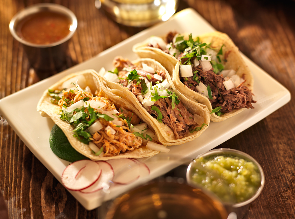 A plate with three different types of tacos on it. They are shredded chicken, shredded pork, and shredded beef. They have fresh cut cilantro and onion sprinkled on top. There is a lime wedge on the plate and dishes of different salsas on the wood table around the plate. A great meal to be found at restaurants in Austin. 