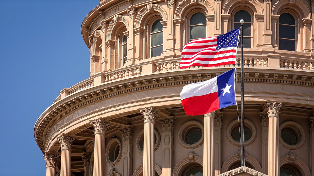 An ornate state building in Texas. It is made of a tan colored stone and has lots of intricate carvings and details. In front of it there is an American and Texas flag flying on a flag pole. 