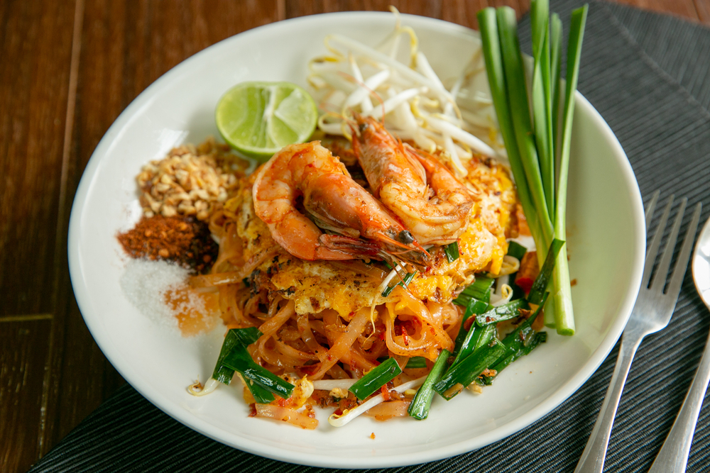 A white bowl on a wooden table and greyish blue table cloth. In the bowl there is a Thai noodle dish. It has green onion, large shrimp, a slice of lime, bamboo shoots, and red pepper flakes. Its similar to what you can find at restaurants in Amarillo. 