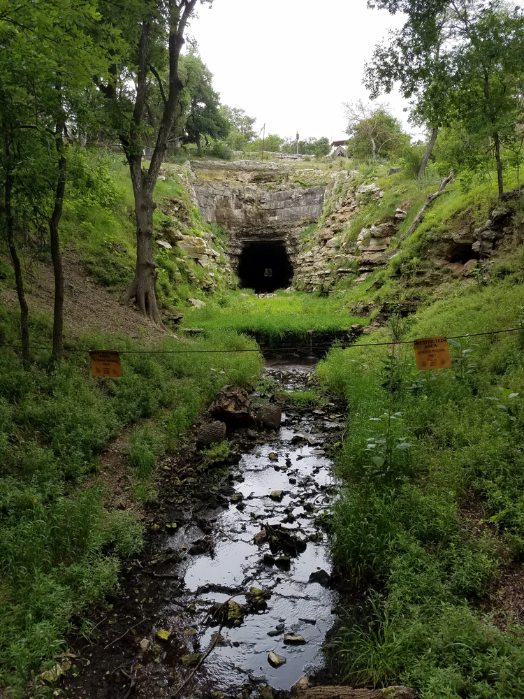 A dark old tunnel roped off by caution signs with a shallow creek in the foreground