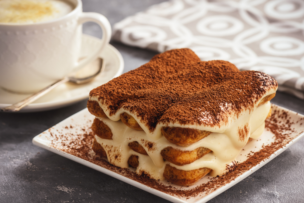 A close up of a small dessert plate with a slice of Tiramisu on it. You can see the layers of sponge cake, the creamy filling flowing over the sides, and it is covered in chocolate powder. Behind the plate you can see a white cup of coffee with a spoon on the saucer. A delish dessert option at one of the restaurants in Austin. 