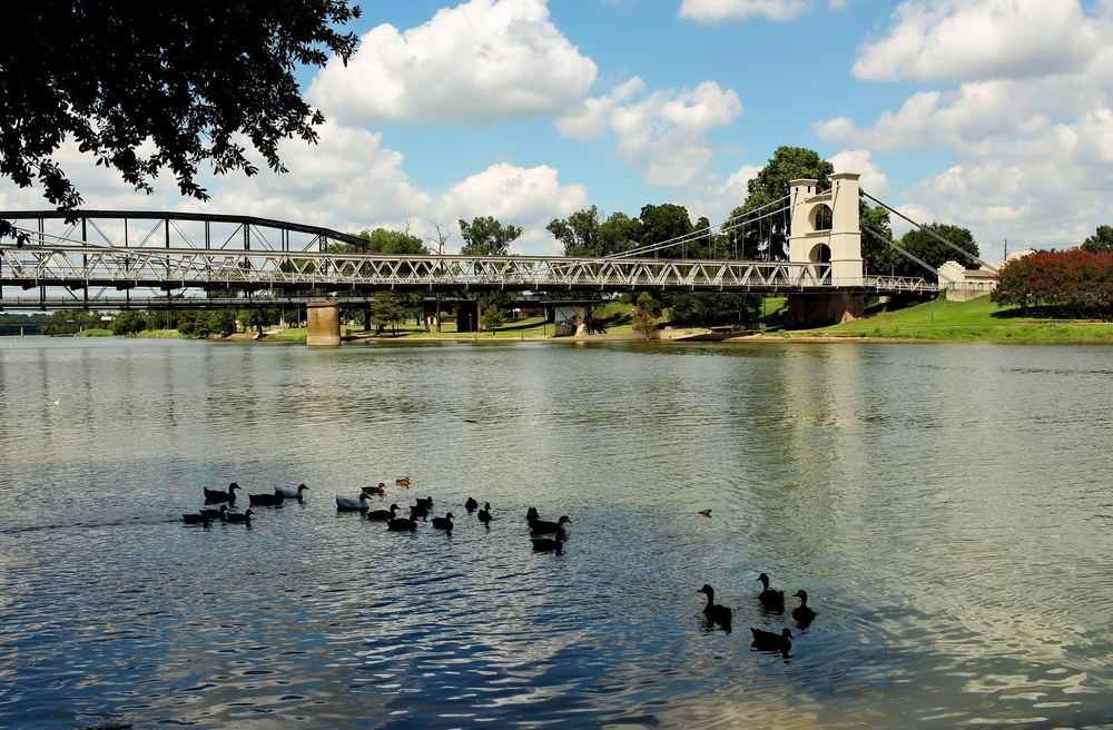 A view of the river and bridge in Waco. The bridge has a white tower like entrance and there are ducks on the river. There is a grassy area with some trees near the bridge. It is a sunny day with big fluffy clouds. One of the best day trips from Dallas. 