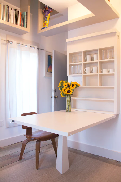 a small white Bright room with white mugs and glasses on a shelf over a table with Sunflowers in a vase on it. 