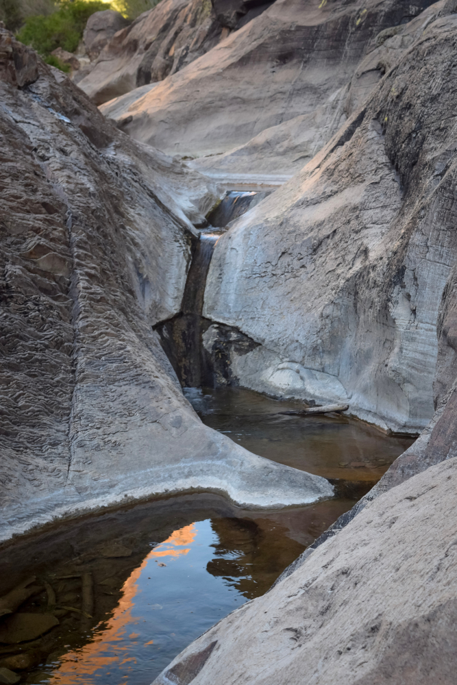 A thin waterfall dropping over several layers of rock into a pool that continues flowing. The rocks are gray and sand in color. You can see where the water has carved into the rock over time.