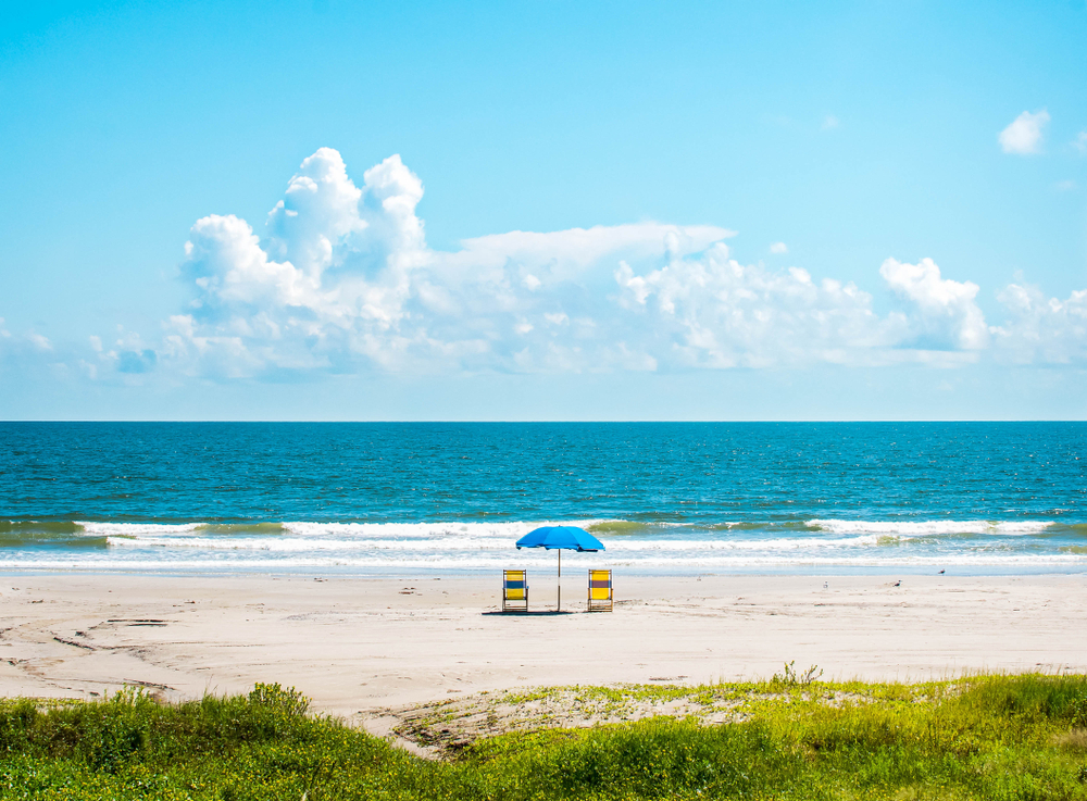 a lone blue umbrella with 2 yellow beach chairs on either side of it facing the ocean on a sunny day at one of the best beaches in texas