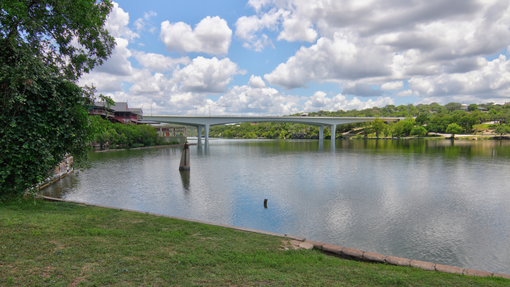 long silver bridge crossing over a lake surrounded by trees on a bright sunny day with blue skies and clouds