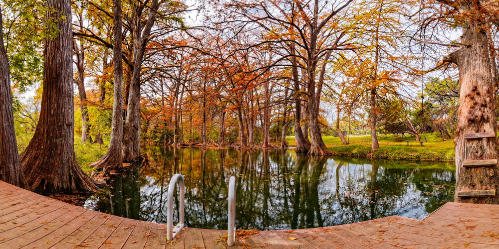 Red dock over looks a large waterway of clear water. The banks on each side are bright green with trees whose leaves are turning orange.